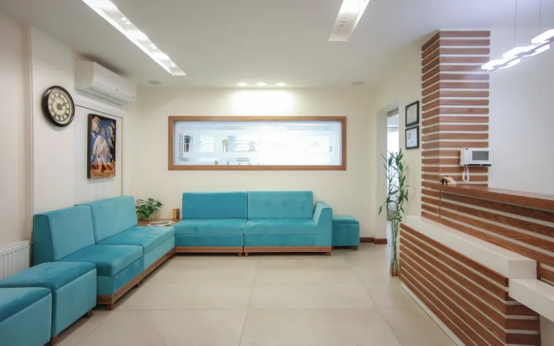 A clinic waiting room with teal sofas, a wall clock and a wood slatted reception desk.