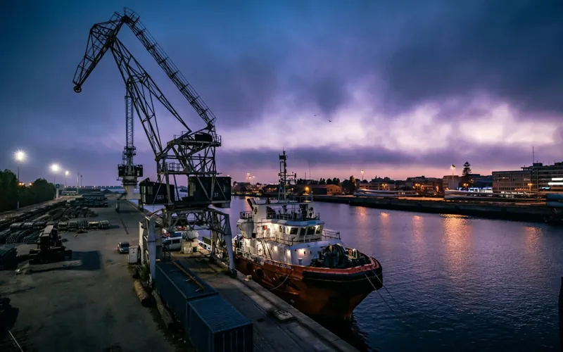 A harbour crane and a tugboat at a quayside at dusk under a purple sky.