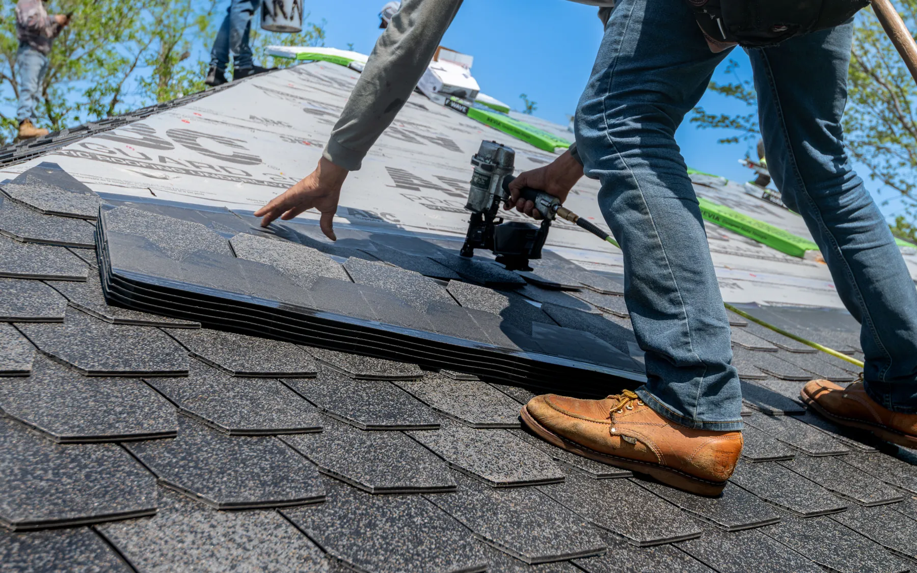 A roofer kneels on a half-finished shingle roof and drives nails with a nail gun, underlayment showing beside him.