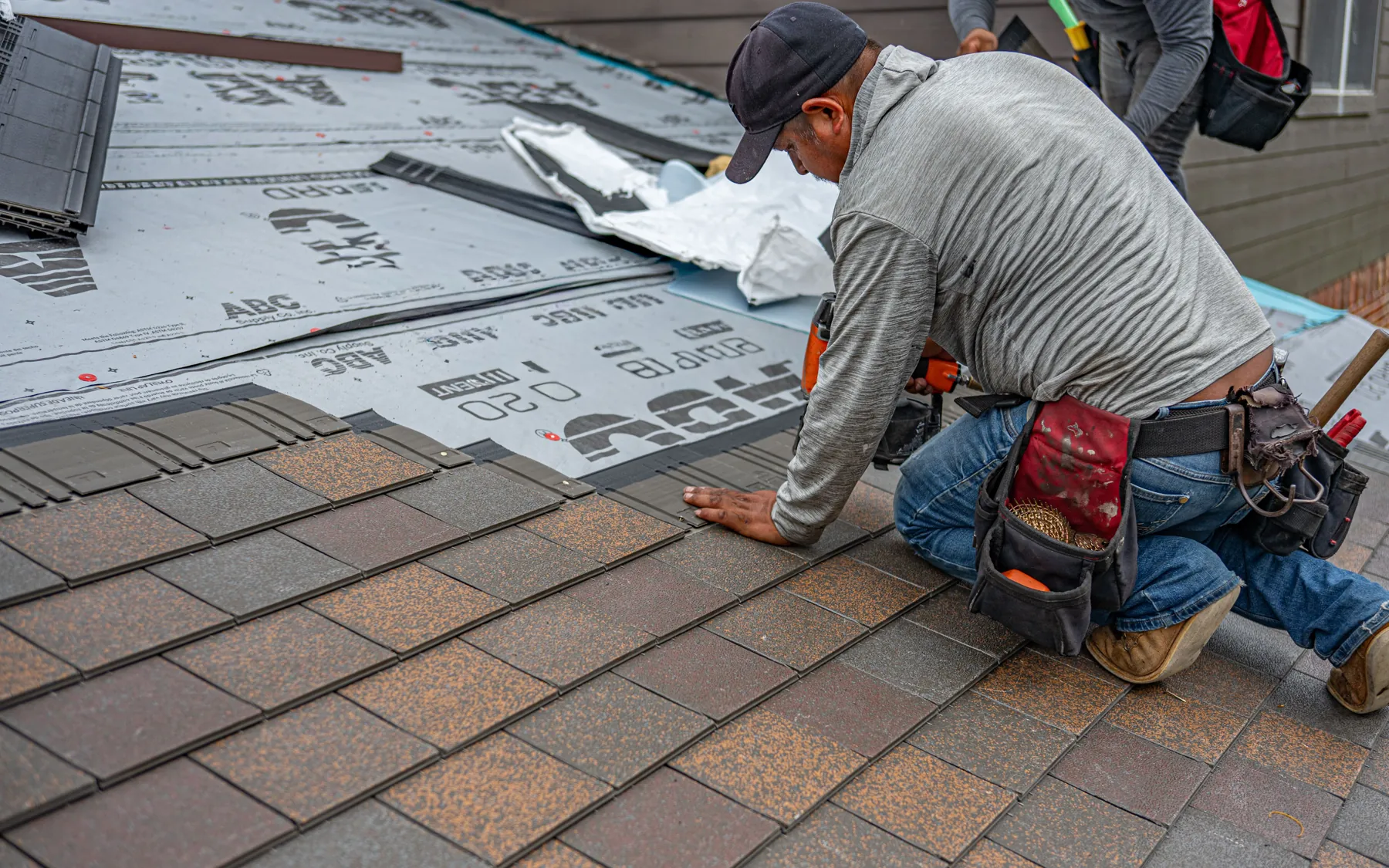 A roofer in a cap and tool belt kneels on a roof deck, nailing down new asphalt shingles over fresh underlayment.