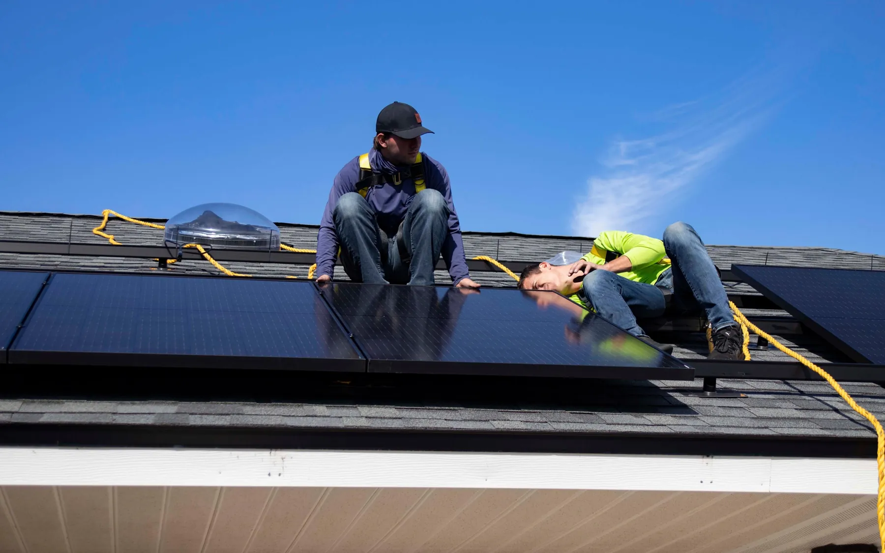 Two installers kneel on a shingled house roof, fastening a row of black solar panels under a clear sky.